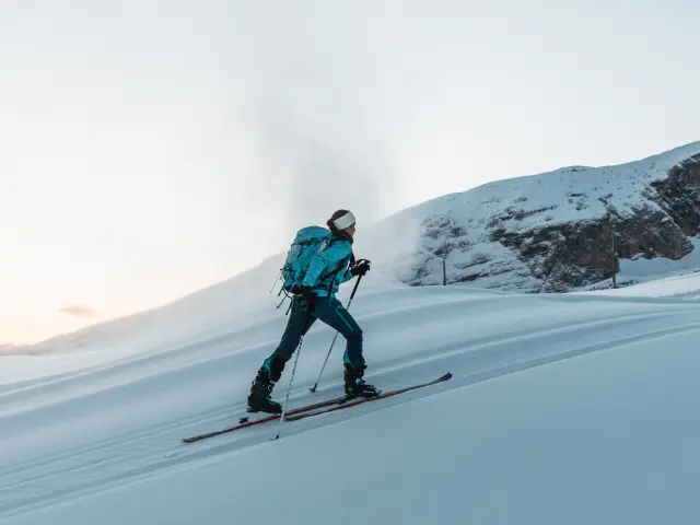Personne en ski de randonnée montant une pente enneigée avec un sac à dos, lumière douce d’hiver.