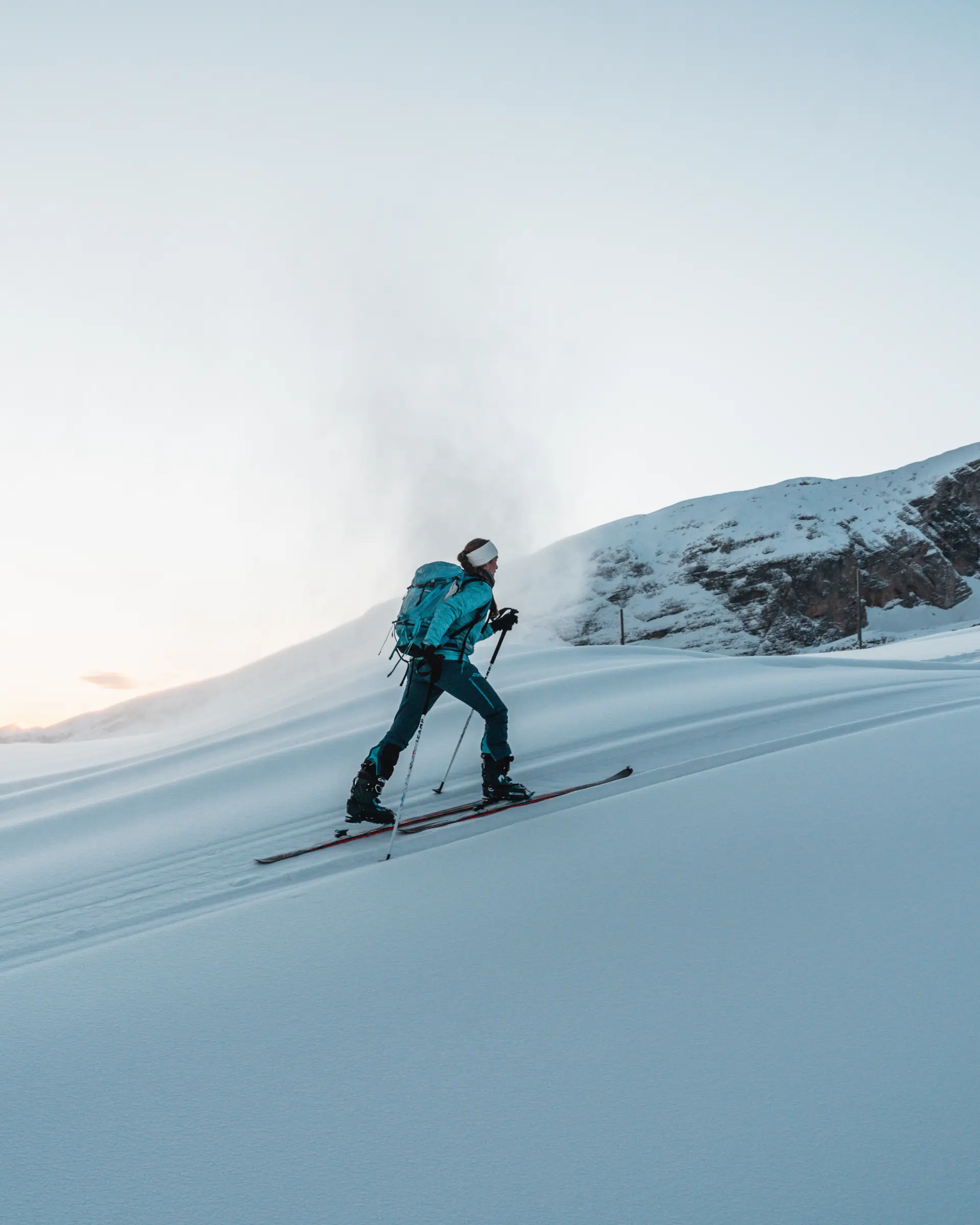 Personne en ski de randonnée montant une pente enneigée avec un sac à dos, lumière douce d’hiver.