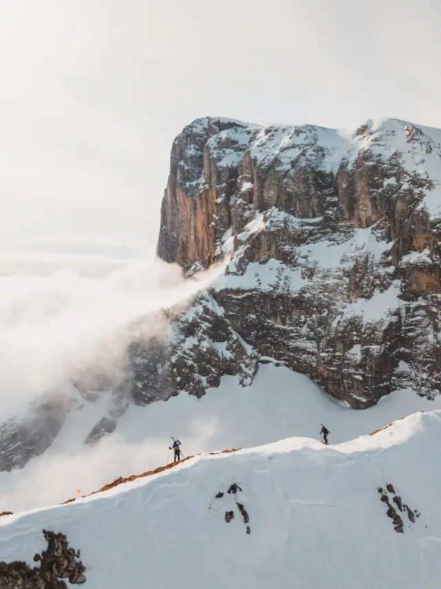 Two ski mountaineers walking along a narrow snowy ridge during La Grande Trace Winter, with limestone cliffs and clouds below.