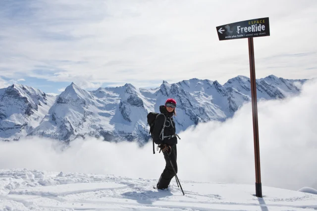 A smiling skier poses in front of the Espace Freeride sign with a panoramic view of the Aravis mountain range and a growing cloud of powder behind her. She is dressed in a black ski suit with a rucksack, mask, poles and a fuchsia pink woollen hat.