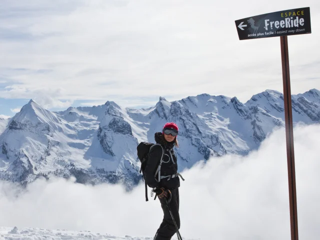 Une skieuse souriante pose devant la pancarte de l'Espace Freeride avec une vue panoramique sur la chaîne des Aravis et un nuage de poudreuse grandissant derrière la skieuse. Elle est vêtue d'un ensemble de ski noir avec un sac à dos, un masque, des bâtons et un bonnet en laine rose fuchsia.