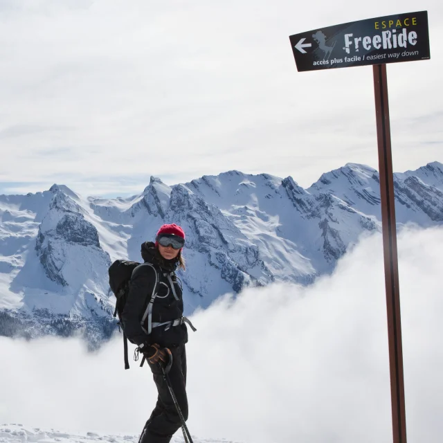 Une skieuse souriante pose devant la pancarte de l'Espace Freeride avec une vue panoramique sur la chaîne des Aravis et un nuage de poudreuse grandissant derrière la skieuse. Elle est vêtue d'un ensemble de ski noir avec un sac à dos, un masque, des bâtons et un bonnet en laine rose fuchsia.