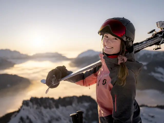 Une jeune femme se tient face à un paysage enneigé, le mont Bagy, au coucher du soleil. Elle porte des vêtements roses et gris et des équipements de ski. Elle porte des skis sur son épaule et son expression est joyeuse, avec un large sourire dirigé vers l'appareil photo. L'image indique un moment de bonheur et de joie au couché du soleil avec une vue panoramique à couper le souffle.