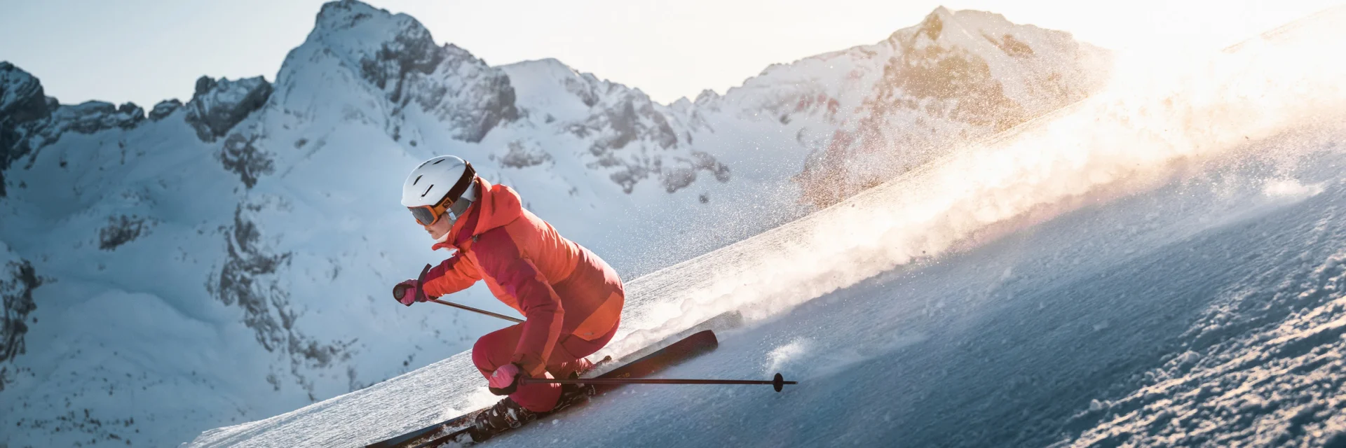 Alpine skier descending a slope with a panoramic view of the Aravis mountain range in the background. The sky is fairly clear, with a lovely golden glow moving behind the skier, wearing a pink ski suit.