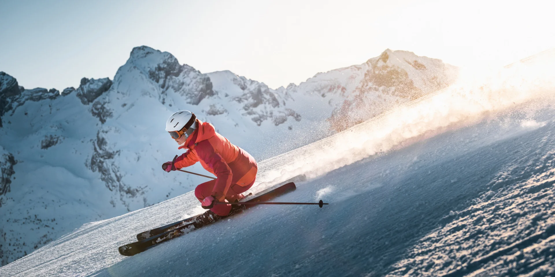 Skieuse alpine qui dévale une pente avec une vue panoramique sur la chaîne des Aravis en arrière-plan. Le ciel est plutôt clair avec une belle luminosité dorée qui vient se déplacer derrière la skieuse, vêtue d'un ensemble de ski en nuances de rose.