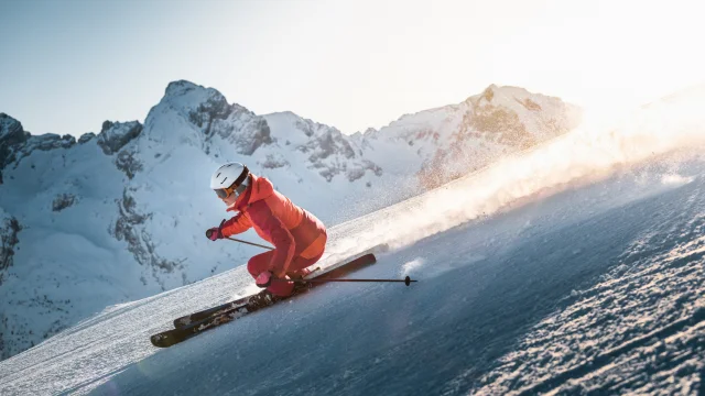Alpine skier descending a slope with a panoramic view of the Aravis mountain range in the background. The sky is fairly clear, with a lovely golden glow moving behind the skier, wearing a pink ski suit.