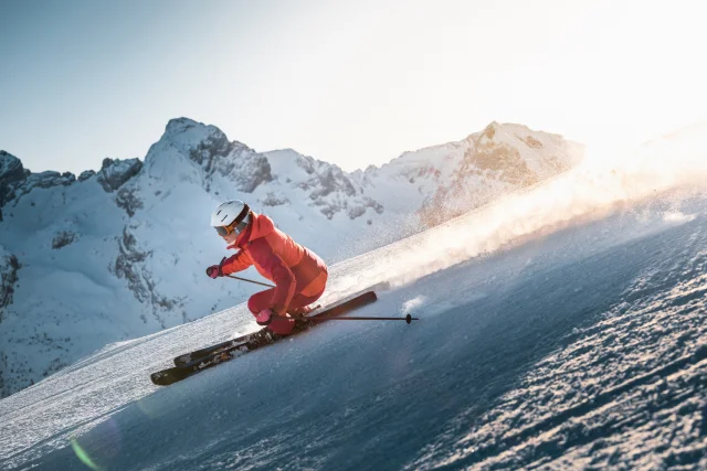 Alpine skier descending a slope with a panoramic view of the Aravis mountain range in the background. The sky is fairly clear, with a lovely golden glow moving behind the skier, wearing a pink ski suit.