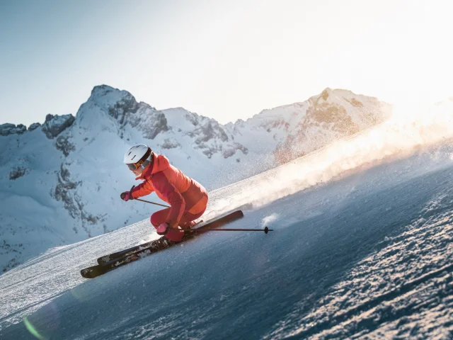 Skieuse alpine qui dévale une pente avec une vue panoramique sur la chaîne des Aravis en arrière-plan. Le ciel est plutôt clair avec une belle luminosité dorée qui vient se déplacer derrière la skieuse, vêtue d'un ensemble de ski en nuances de rose.