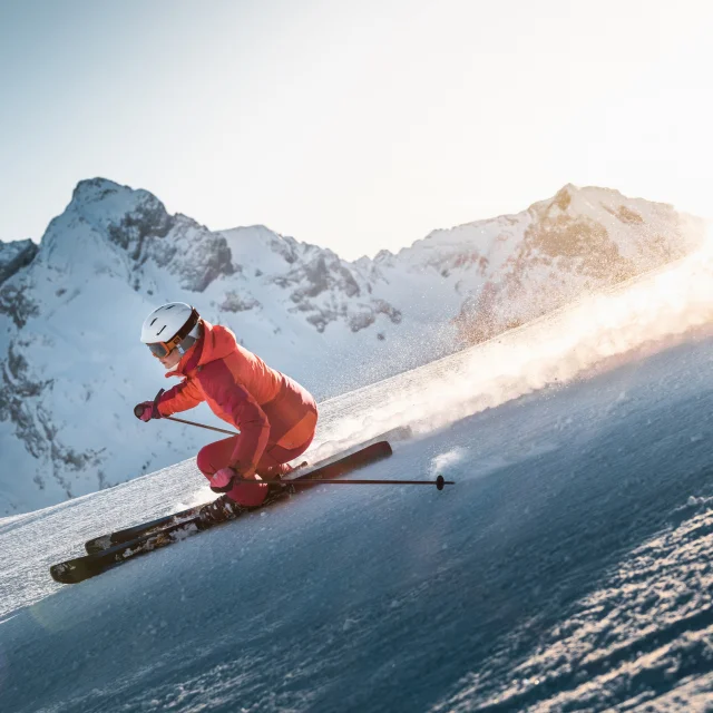Alpine skier descending a slope with a panoramic view of the Aravis mountain range in the background. The sky is fairly clear, with a lovely golden glow moving behind the skier, wearing a pink ski suit.