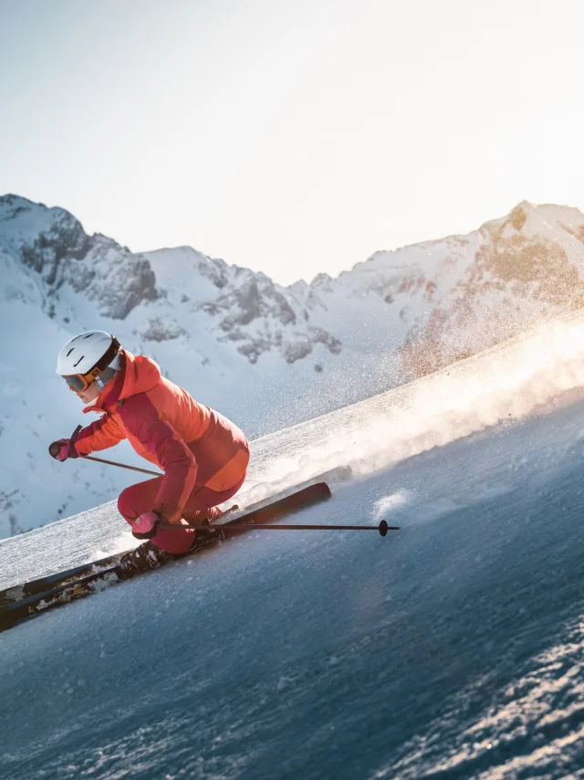 Skieuse alpine qui dévale une pente avec une vue panoramique sur la chaîne des Aravis en arrière-plan. Le ciel est plutôt clair avec une belle luminosité dorée qui vient se déplacer derrière la skieuse, vêtue d'un ensemble de ski en nuances de rose.