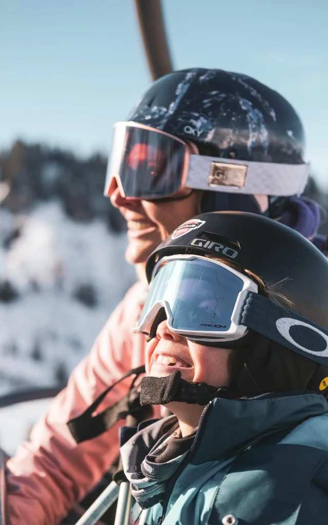 Eine Mutter und ihr Sohn befinden sich in einem Sessellift, der sie auf die Skipiste hinaufbringt. Die beiden sind sehr vertraut und scheinen sich auf das Skifahren zu freuen, wenn man ihr breites Lächeln und ihren Gesichtsausdruck betrachtet, während sie die Skipiste betrachten.