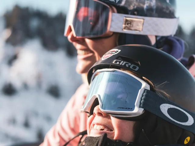 Une mère et son fils sont sur un télésiège pour monter en haut de la piste de ski. Ils sont tous deux très complices et semblent être impatients de skier, au vu de leur grand sourire et expression du visage, contemplant les pistes de ski.