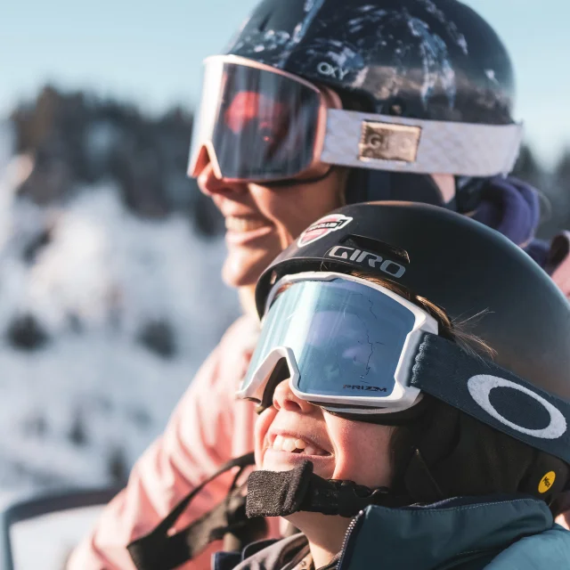 Une mère et son fils sont sur un télésiège pour monter en haut de la piste de ski. Ils sont tous deux très complices et semblent être impatients de skier, au vu de leur grand sourire et expression du visage, contemplant les pistes de ski.