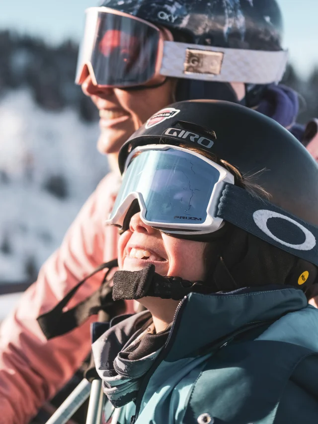 Une mère et son fils sont sur un télésiège pour monter en haut de la piste de ski. Ils sont tous deux très complices et semblent être impatients de skier, au vu de leur grand sourire et expression du visage, contemplant les pistes de ski.