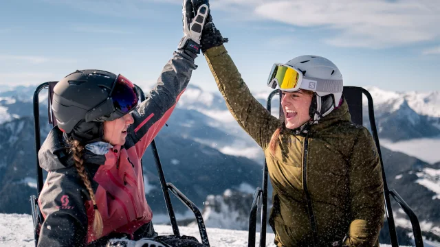 2 amies en habits de ski, sont assises sur des chaises longues en haut d'une montagne enneigée, elles se tapent dans la main avec un grand sourire. Il y a une vue panoramique sur la chaîne des Aravis en arrière-plan. Elles profitent d'un moment de détente sous un ciel ensoleillé. Une personne porte une veste rose, une autre une verte. L'ambiance est conviviale et détendue, évoquant une pause après une séance de ski.