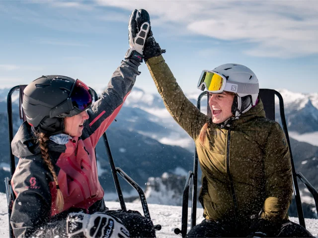 2 amies en habits de ski, sont assises sur des chaises longues en haut d'une montagne enneigée, elles se tapent dans la main avec un grand sourire. Il y a une vue panoramique sur la chaîne des Aravis en arrière-plan. Elles profitent d'un moment de détente sous un ciel ensoleillé. Une personne porte une veste rose, une autre une verte. L'ambiance est conviviale et détendue, évoquant une pause après une séance de ski.