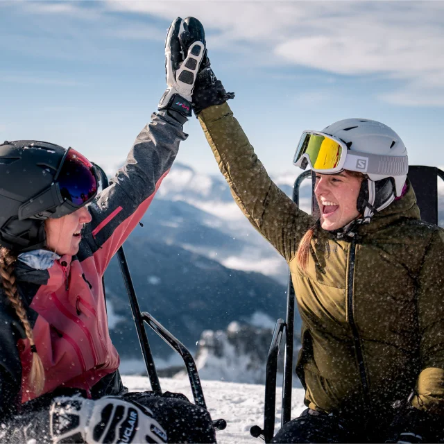 2 amies en habits de ski, sont assises sur des chaises longues en haut d'une montagne enneigée, elles se tapent dans la main avec un grand sourire. Il y a une vue panoramique sur la chaîne des Aravis en arrière-plan. Elles profitent d'un moment de détente sous un ciel ensoleillé. Une personne porte une veste rose, une autre une verte. L'ambiance est conviviale et détendue, évoquant une pause après une séance de ski.