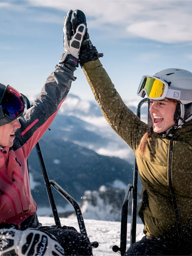 2 amies en habits de ski, sont assises sur des chaises longues en haut d'une montagne enneigée, elles se tapent dans la main avec un grand sourire. Il y a une vue panoramique sur la chaîne des Aravis en arrière-plan. Elles profitent d'un moment de détente sous un ciel ensoleillé. Une personne porte une veste rose, une autre une verte. L'ambiance est conviviale et détendue, évoquant une pause après une séance de ski.