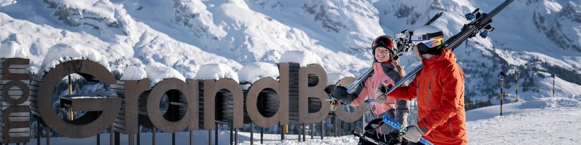 Deux personnes en tenue de ski, portant des skis sur l'épaule, marchent sur une piste enneigée avec un large sourire. En arrière-plan, on peut voir un grand panneau en bois indiquant mon GrandBo, ainsi que la chaîne des Aravis enneigée sous un ciel bleu. L'atmosphère est joyeuse et décontractée.
