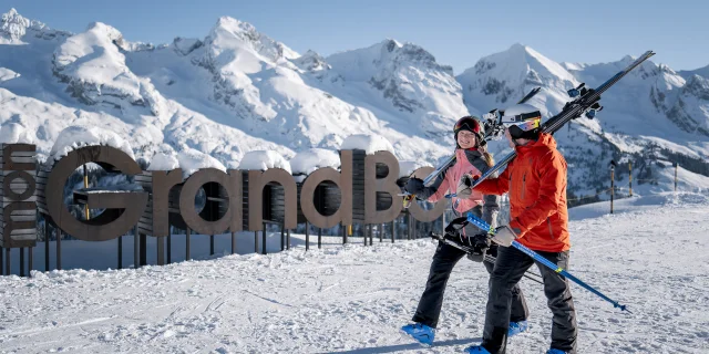 Deux personnes en tenue de ski, portant des skis sur l'épaule, marchent sur une piste enneigée avec un large sourire. En arrière-plan, on peut voir un grand panneau en bois indiquant mon GrandBo, ainsi que la chaîne des Aravis enneigée sous un ciel bleu. L'atmosphère est joyeuse et décontractée.