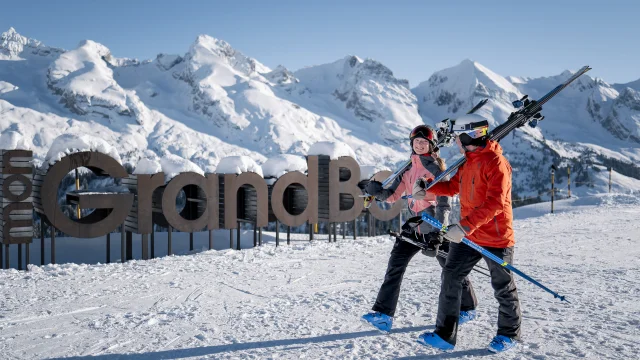 Two people in ski outfits, with skis slung over their shoulders, walk down a snow-covered slope with broad smiles on their faces. In the background, you can see a large wooden sign reading mon GrandBo, as well as the Aravis mountain range covered in snow under a blue sky. The atmosphere is happy and relaxed.