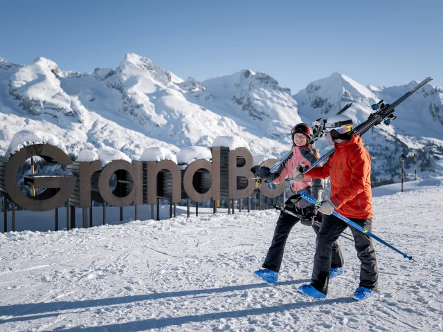 Two people in ski outfits, with skis slung over their shoulders, walk down a snow-covered slope with broad smiles on their faces. In the background, you can see a large wooden sign reading mon GrandBo, as well as the Aravis mountain range covered in snow under a blue sky. The atmosphere is happy and relaxed.