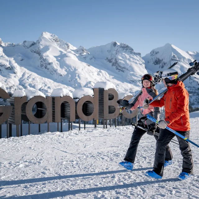 Deux personnes en tenue de ski, portant des skis sur l'épaule, marchent sur une piste enneigée avec un large sourire. En arrière-plan, on peut voir un grand panneau en bois indiquant mon GrandBo, ainsi que la chaîne des Aravis enneigée sous un ciel bleu. L'atmosphère est joyeuse et décontractée.