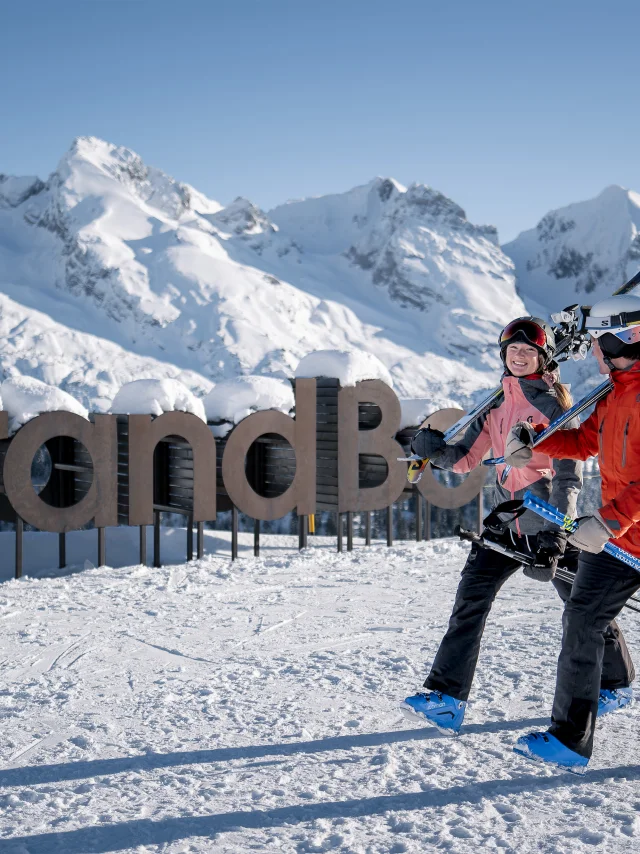 Deux personnes en tenue de ski, portant des skis sur l'épaule, marchent sur une piste enneigée avec un large sourire. En arrière-plan, on peut voir un grand panneau en bois indiquant mon GrandBo, ainsi que la chaîne des Aravis enneigée sous un ciel bleu. L'atmosphère est joyeuse et décontractée.