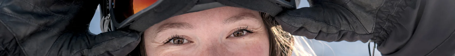 Gros plan d'une jeune femme souriante, portant une veste de ski rose et grise, des gants noirs, et un casque avec des lunettes de ski. Elle ajuste ses lunettes avec ses mains. En arrière-plan, on distingue des montagnes enneigées sous un ciel bleu. Son expression rayonnante et le cadre alpin donnent une impression de joie et de plaisir en montagne.