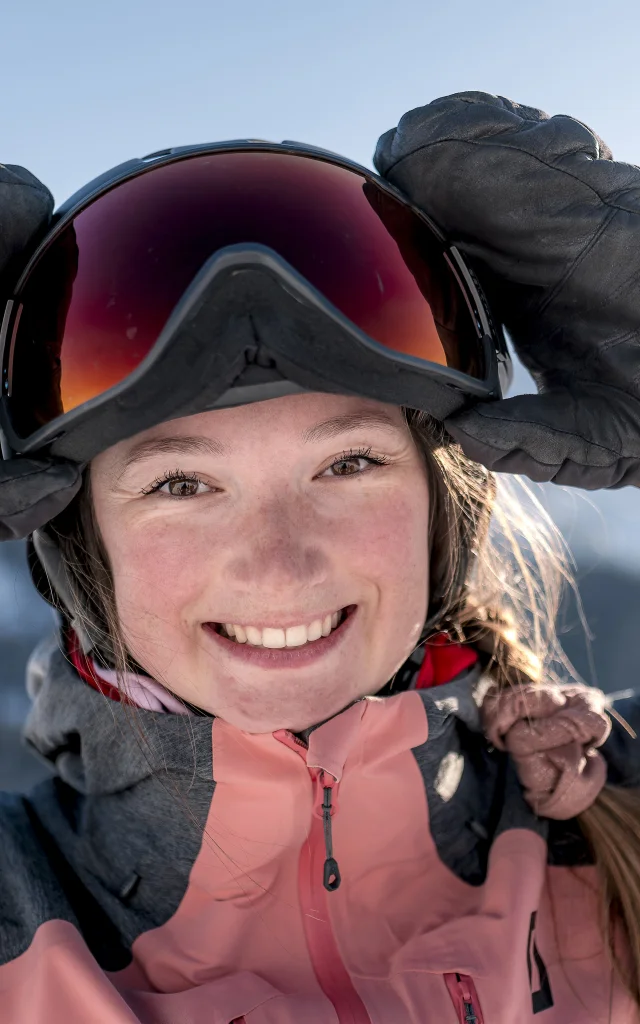 Nahaufnahme einer lächelnden jungen Frau, die eine rosa-graue Skijacke, schwarze Handschuhe und einen Helm mit einer Skibrille trägt. Sie rückt die Brille mit den Händen zurecht. Im Hintergrund sind schneebedeckte Berge unter einem blauen Himmel zu erkennen. Ihr strahlender Gesichtsausdruck und die alpine Umgebung vermitteln den Eindruck von Freude und Spaß in den Bergen.