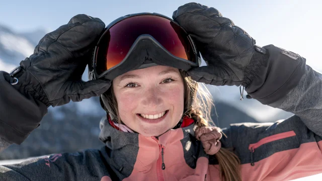 Gros plan d'une jeune femme souriante, portant une veste de ski rose et grise, des gants noirs, et un casque avec des lunettes de ski. Elle ajuste ses lunettes avec ses mains. En arrière-plan, on distingue des montagnes enneigées sous un ciel bleu. Son expression rayonnante et le cadre alpin donnent une impression de joie et de plaisir en montagne.
