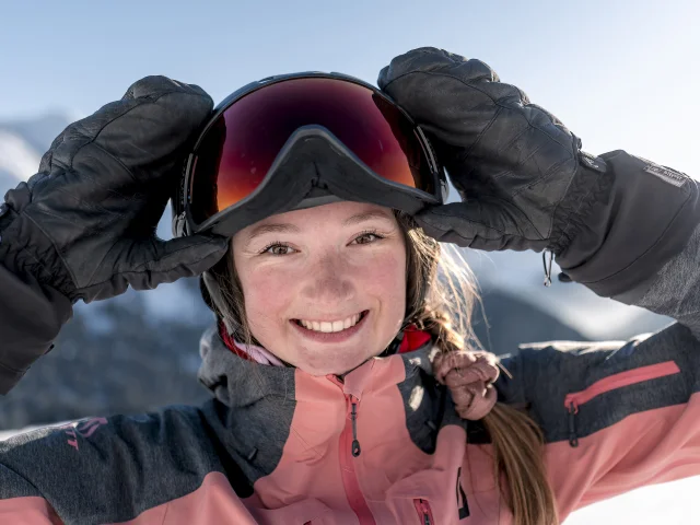 Close-up of a smiling young woman wearing a pink and grey ski jacket, black gloves and a helmet with ski goggles. She is adjusting her glasses with her hands. In the background are snow-capped mountains under a blue sky. Her radiant expression and the alpine setting give an impression of joy and pleasure in the mountains.