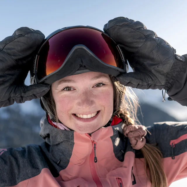 Nahaufnahme einer lächelnden jungen Frau, die eine rosa-graue Skijacke, schwarze Handschuhe und einen Helm mit einer Skibrille trägt. Sie rückt die Brille mit den Händen zurecht. Im Hintergrund sind schneebedeckte Berge unter einem blauen Himmel zu erkennen. Ihr strahlender Gesichtsausdruck und die alpine Umgebung vermitteln den Eindruck von Freude und Spaß in den Bergen.