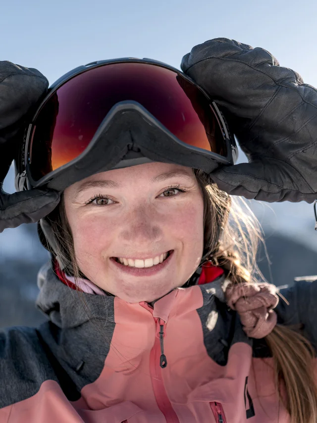Gros plan d'une jeune femme souriante, portant une veste de ski rose et grise, des gants noirs, et un casque avec des lunettes de ski. Elle ajuste ses lunettes avec ses mains. En arrière-plan, on distingue des montagnes enneigées sous un ciel bleu. Son expression rayonnante et le cadre alpin donnent une impression de joie et de plaisir en montagne.