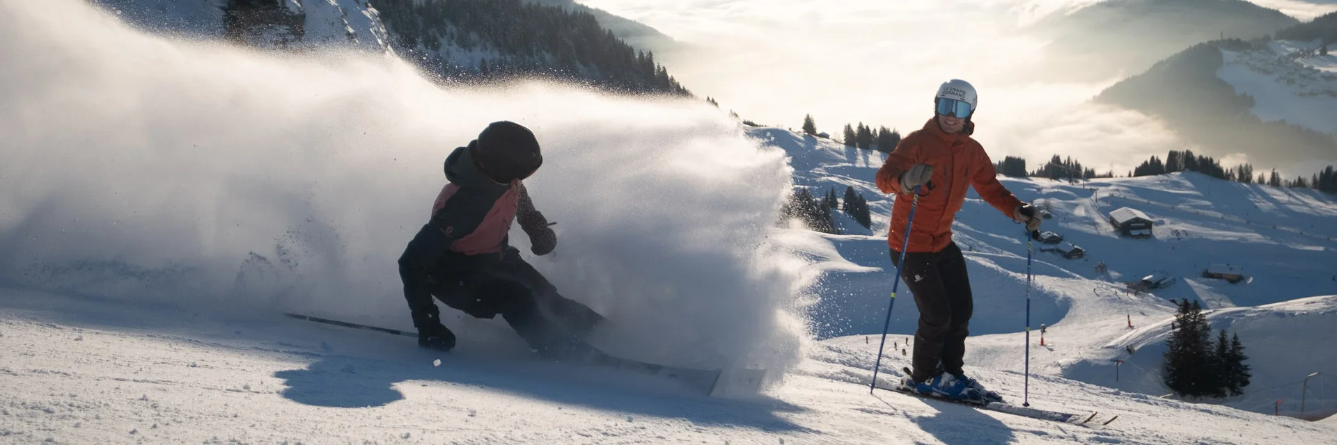 Deux skieurs sur une piste enneigée en pleine descente. L'un des skieurs, vêtu d'une veste rouge, se tient debout en arrière-plan, tandis que l'autre, en train de skier rapidement en avant-plan, soulève un nuage de poudreuse impressionnant. En arrière-plan, on peut voir le massif du Bargy avec une mer de nuages couvrant la vallée, le tout sous un ciel bleu ensoleillé. L'ambiance évoque l'aventure, la liberté et la joie.