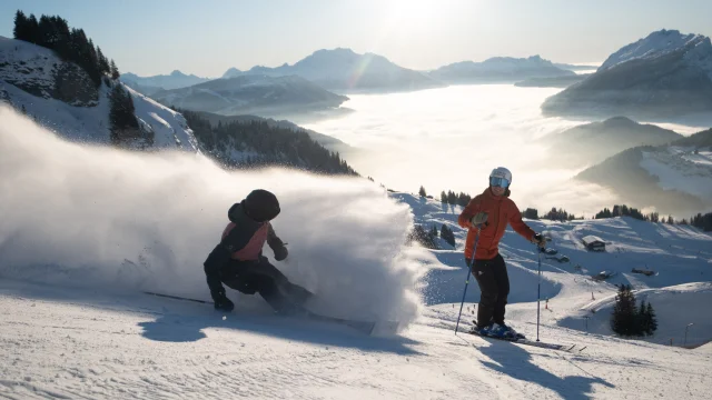 Deux skieurs sur une piste enneigée en pleine descente. L'un des skieurs, vêtu d'une veste rouge, se tient debout en arrière-plan, tandis que l'autre, en train de skier rapidement en avant-plan, soulève un nuage de poudreuse impressionnant. En arrière-plan, on peut voir le massif du Bargy avec une mer de nuages couvrant la vallée, le tout sous un ciel bleu ensoleillé. L'ambiance évoque l'aventure, la liberté et la joie.