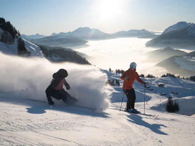 Deux skieurs sur une piste enneigée en pleine descente. L'un des skieurs, vêtu d'une veste rouge, se tient debout en arrière-plan, tandis que l'autre, en train de skier rapidement en avant-plan, soulève un nuage de poudreuse impressionnant. En arrière-plan, on peut voir le massif du Bargy avec une mer de nuages couvrant la vallée, le tout sous un ciel bleu ensoleillé. L'ambiance évoque l'aventure, la liberté et la joie.
