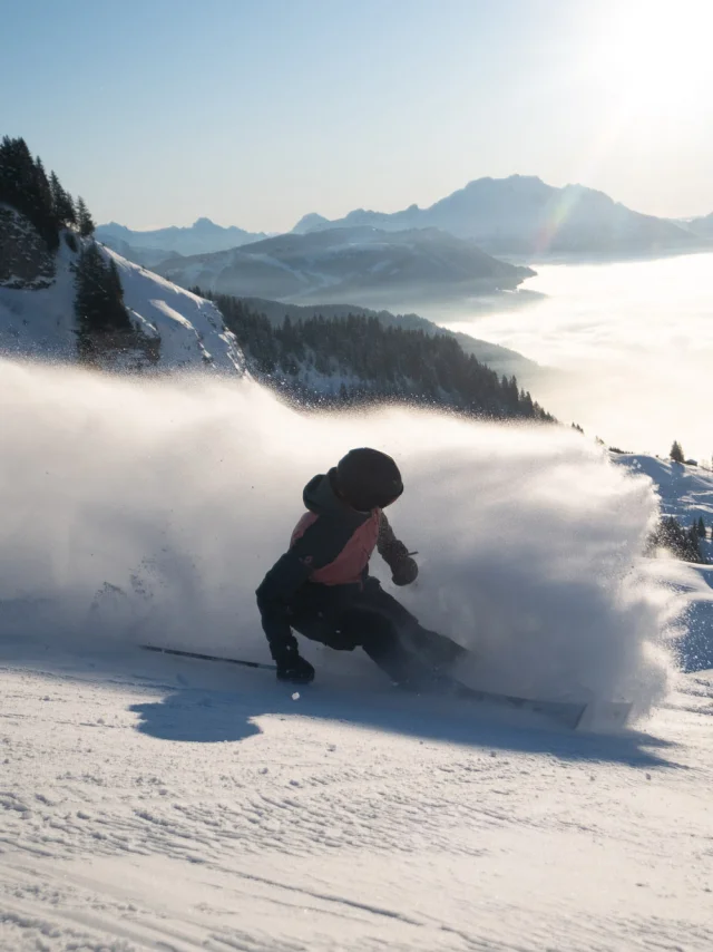 Deux skieurs sur une piste enneigée en pleine descente. L'un des skieurs, vêtu d'une veste rouge, se tient debout en arrière-plan, tandis que l'autre, en train de skier rapidement en avant-plan, soulève un nuage de poudreuse impressionnant. En arrière-plan, on peut voir le massif du Bargy avec une mer de nuages couvrant la vallée, le tout sous un ciel bleu ensoleillé. L'ambiance évoque l'aventure, la liberté et la joie.