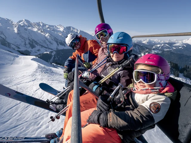 Une famille sur un télésiège entourée par la chaîne des Aravis enneigée sous un ciel dégagé. Tous portent des casques de ski colorés (rose, bleu, violet et blanc) ainsi que des lunettes de ski. En arrière-plan, on peut voir des pistes de ski et des arbres enneigés ce qui indique une belle journée ensoleillée en montagne. L'ambiance générale est celle d'un moment convivial en famille en plein air.