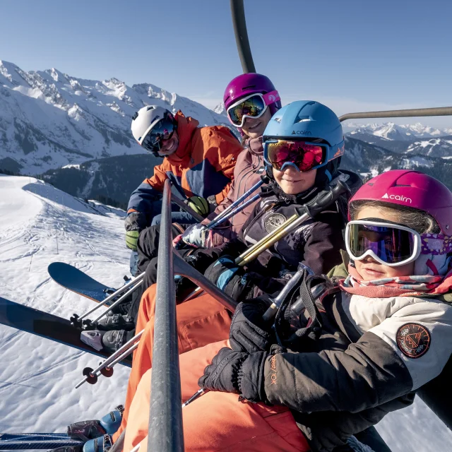 Une famille sur un télésiège entourée par la chaîne des Aravis enneigée sous un ciel dégagé. Tous portent des casques de ski colorés (rose, bleu, violet et blanc) ainsi que des lunettes de ski. En arrière-plan, on peut voir des pistes de ski et des arbres enneigés ce qui indique une belle journée ensoleillée en montagne. L'ambiance générale est celle d'un moment convivial en famille en plein air.