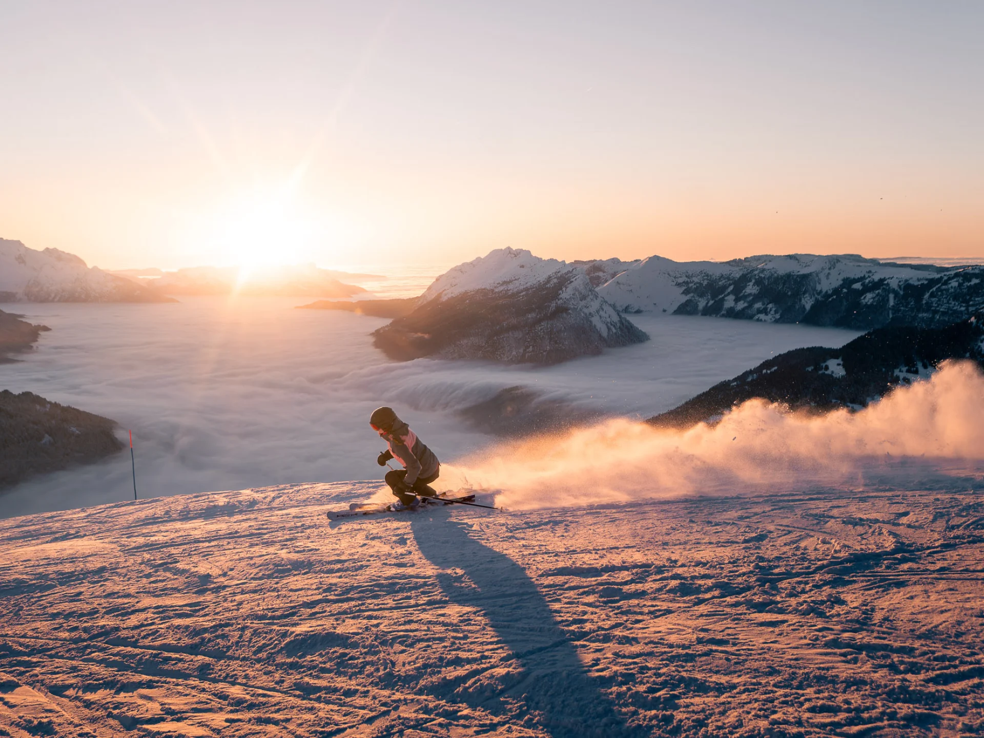 Une skieuse dévale une pente enneigée au coucher du soleil, projetant un nuage de poudreuse derrière elle. En arrière-plan, une mer de nuages enveloppe les vallées, tandis que le mont Bagy se découpe dans une lumière dorée, créant une scène spectaculaire et apaisante.