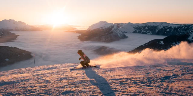 Une skieuse dévale une pente enneigée au coucher du soleil, projetant un nuage de poudreuse derrière elle. En arrière-plan, une mer de nuages enveloppe les vallées, tandis que le mont Bagy se découpe dans une lumière dorée, créant une scène spectaculaire et apaisante.