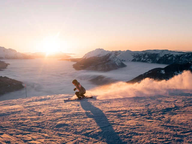 Une skieuse dévale une pente enneigée au coucher du soleil, projetant un nuage de poudreuse derrière elle. En arrière-plan, une mer de nuages enveloppe les vallées, tandis que le mont Bagy se découpe dans une lumière dorée, créant une scène spectaculaire et apaisante.