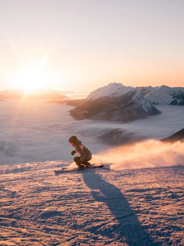 Une skieuse dévale une pente enneigée au coucher du soleil, projetant un nuage de poudreuse derrière elle. En arrière-plan, une mer de nuages enveloppe les vallées, tandis que le mont Bagy se découpe dans une lumière dorée, créant une scène spectaculaire et apaisante.
