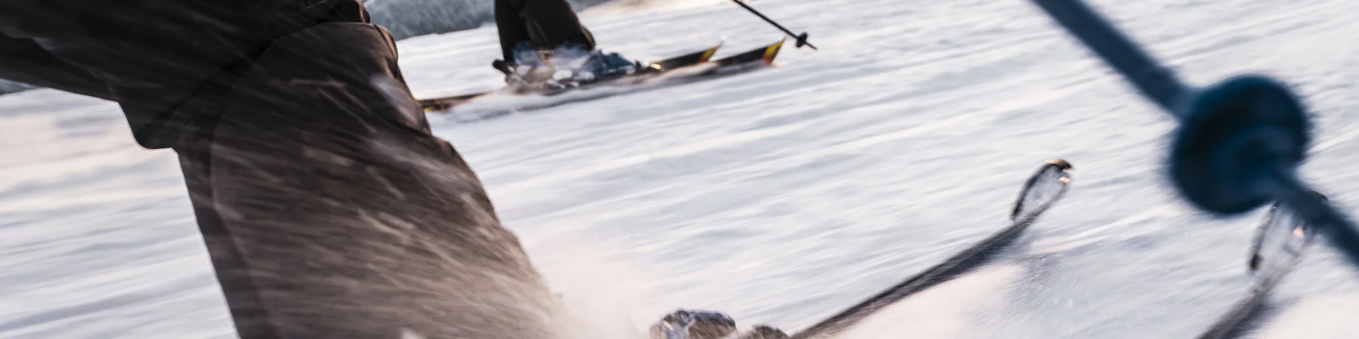 Scène dynamique de skieurs dévalant une piste enneigée. L'accent est mis sur les jambes et les skis du premier skieur, avec des détails nets sur les fixations et la neige projetée par la vitesse. En arrière-plan, un autre skieur, portant une veste rose et noire, glisse en parallèle, avec une vue sur les montagnes et une mer de nuages au loin. La perspective en mouvement donne une sensation d'action et d'énergie.