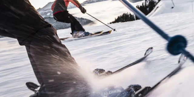 Scène dynamique de skieurs dévalant une piste enneigée. L'accent est mis sur les jambes et les skis du premier skieur, avec des détails nets sur les fixations et la neige projetée par la vitesse. En arrière-plan, un autre skieur, portant une veste rose et noire, glisse en parallèle, avec une vue sur les montagnes et une mer de nuages au loin. La perspective en mouvement donne une sensation d'action et d'énergie.