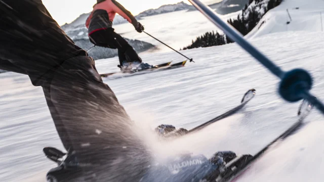 Scène dynamique de skieurs dévalant une piste enneigée. L'accent est mis sur les jambes et les skis du premier skieur, avec des détails nets sur les fixations et la neige projetée par la vitesse. En arrière-plan, un autre skieur, portant une veste rose et noire, glisse en parallèle, avec une vue sur les montagnes et une mer de nuages au loin. La perspective en mouvement donne une sensation d'action et d'énergie.