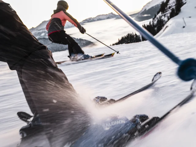 Dynamic scene of skiers hurtling down a snow-covered slope. The focus is on the legs and skis of the first skier, with sharp details of the bindings and the snow thrown up by speed. In the background, another skier, wearing a pink and black jacket, glides along in parallel, with a view of the mountains and a sea of clouds in the distance. The moving perspective gives a sense of action and energy.