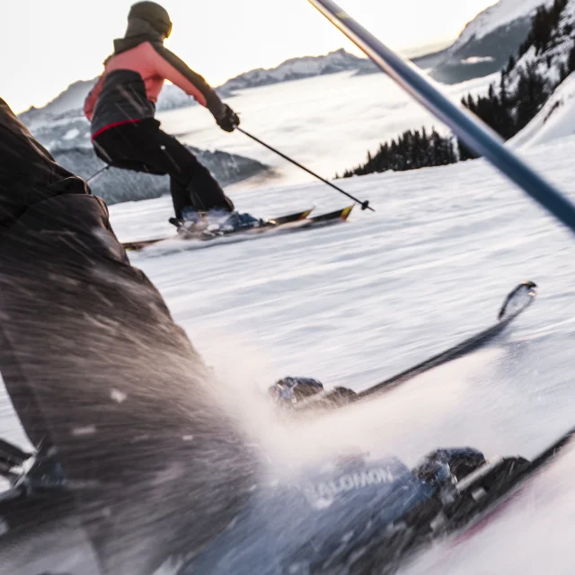 Scène dynamique de skieurs dévalant une piste enneigée. L'accent est mis sur les jambes et les skis du premier skieur, avec des détails nets sur les fixations et la neige projetée par la vitesse. En arrière-plan, un autre skieur, portant une veste rose et noire, glisse en parallèle, avec une vue sur les montagnes et une mer de nuages au loin. La perspective en mouvement donne une sensation d'action et d'énergie.