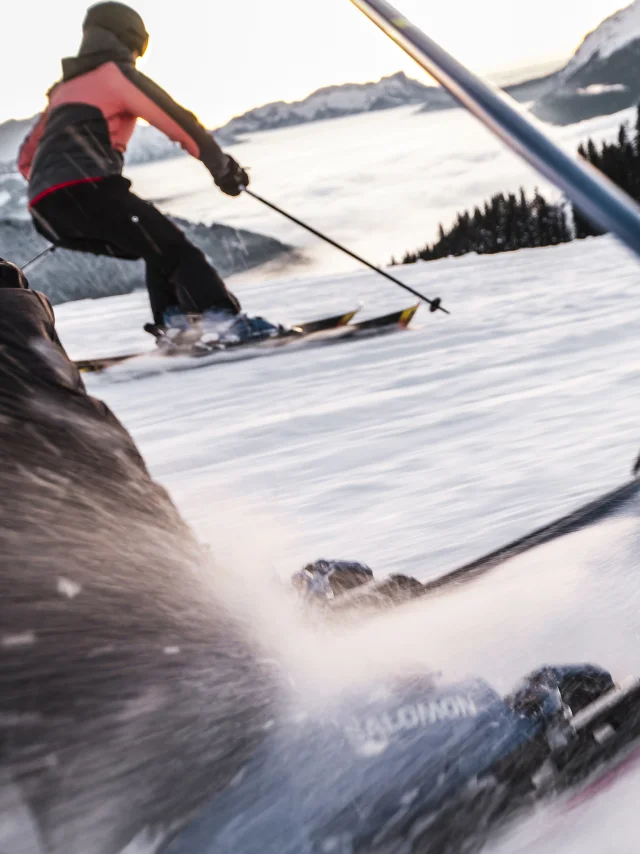 Scène dynamique de skieurs dévalant une piste enneigée. L'accent est mis sur les jambes et les skis du premier skieur, avec des détails nets sur les fixations et la neige projetée par la vitesse. En arrière-plan, un autre skieur, portant une veste rose et noire, glisse en parallèle, avec une vue sur les montagnes et une mer de nuages au loin. La perspective en mouvement donne une sensation d'action et d'énergie.